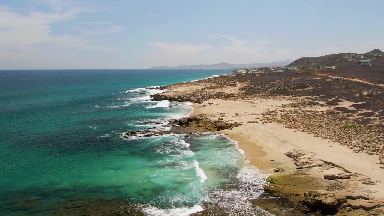 Aerial frame over Buzzards Beach, Baja California Sur, with turquoise shallows, white surf on rocks and sand, dry hillside beyond the shore, and no visible people.