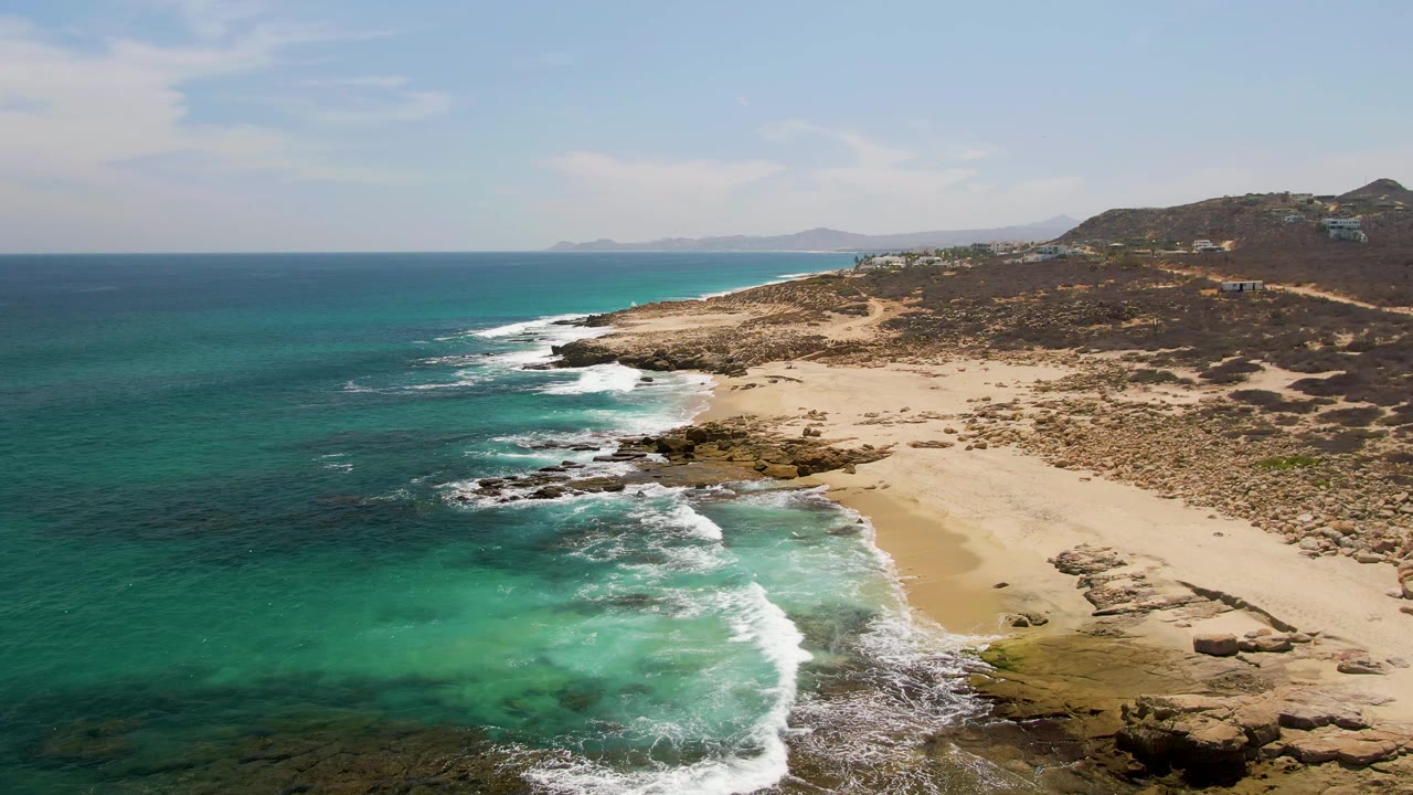 Second frame at Buzzards Beach shows foamy turquoise water advancing across the rocky shallows toward pale sand, with dry scrub hills and no visible people.