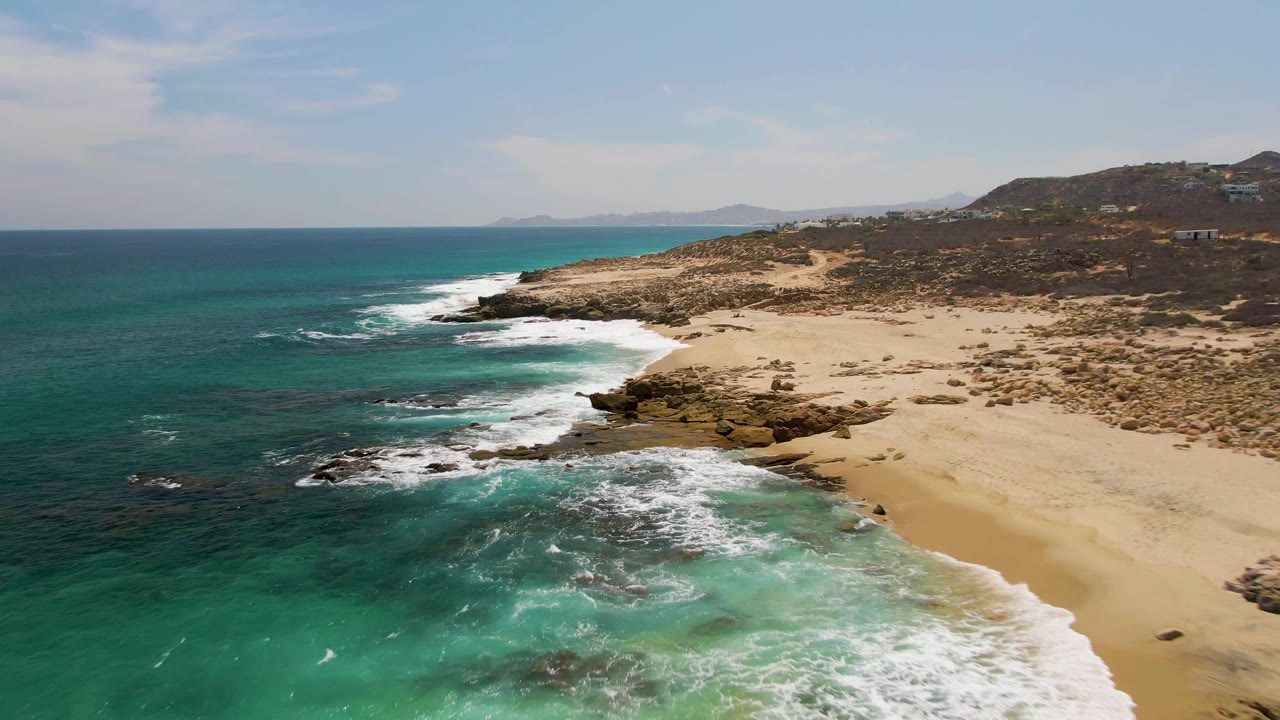 Opening aerial frame over Buzzards Beach in Baja California Sur, with turquoise surf curling onto sand and dark rocks, dry hills in the background, and no visible people.