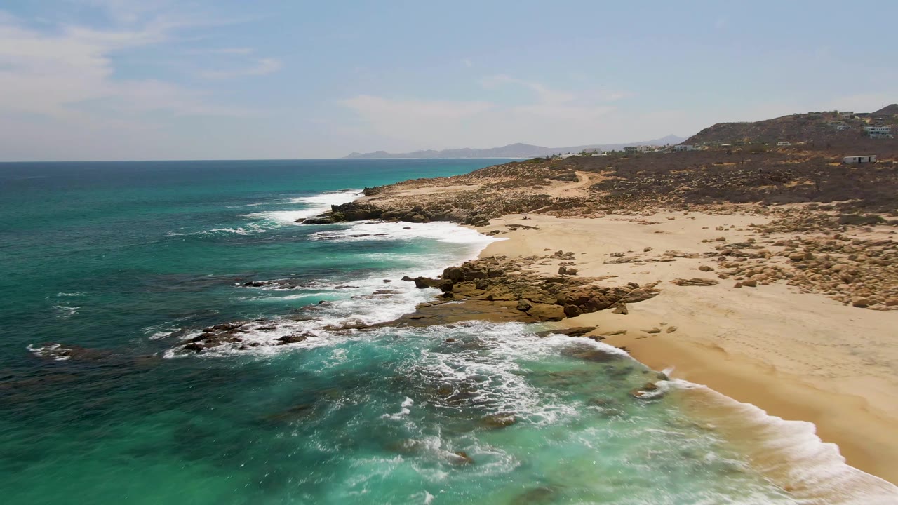 Aerial view at Buzzards Beach with white foam spreading over nearshore rocks, open tan sand to the right, blue-green water to the left, and no visible people.