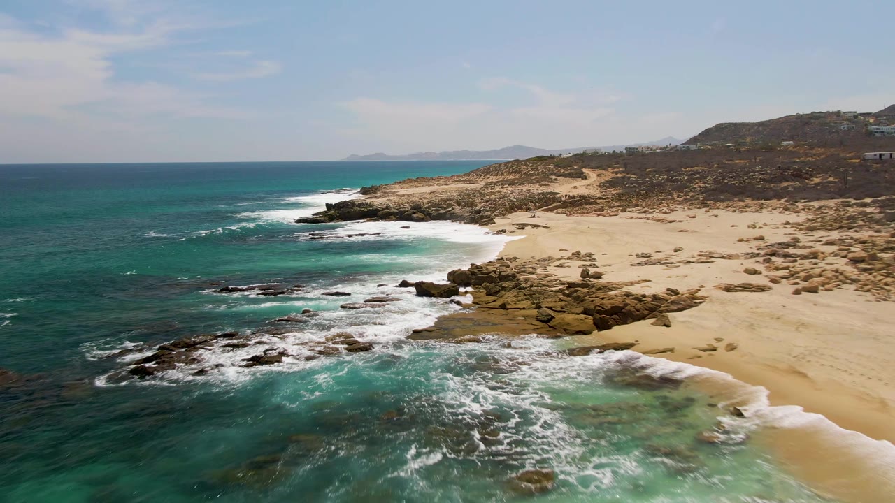 Buzzards Beach coastline seen from above as a fresh band of surf reaches the sand, with exposed rocks in the foreground, low arid hills behind, and no visible people.