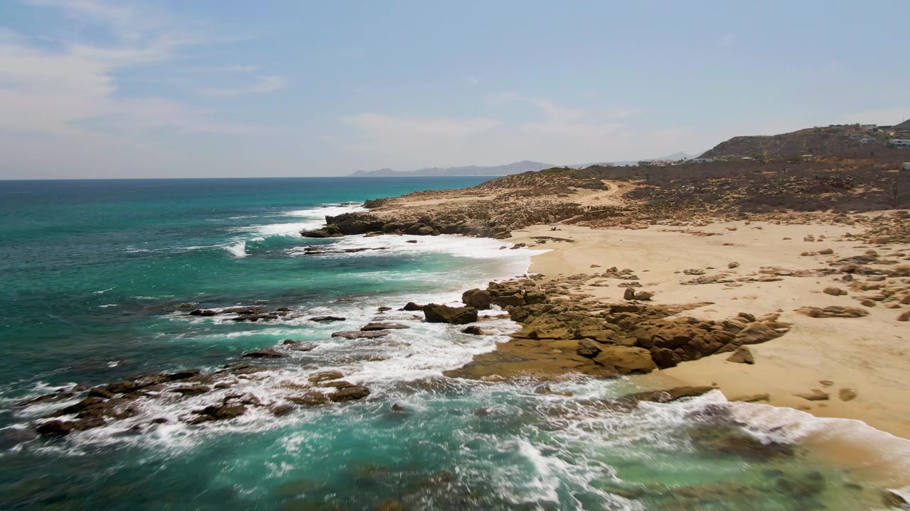 Mid-clip aerial frame over Buzzards Beach, where foam spreads across foreground rocks and shallow green water while the sandy shoreline curves toward distant hills, with no visible people.