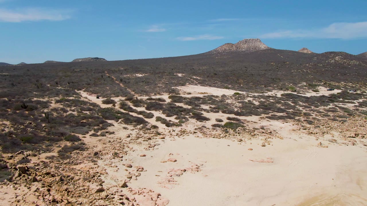 Aerial view at Buzzards Beach, Baja California Sur, showing pale sandy desert, scattered scrub, distant rocky mountains, clear blue sky, and no visible people at the opening moment.