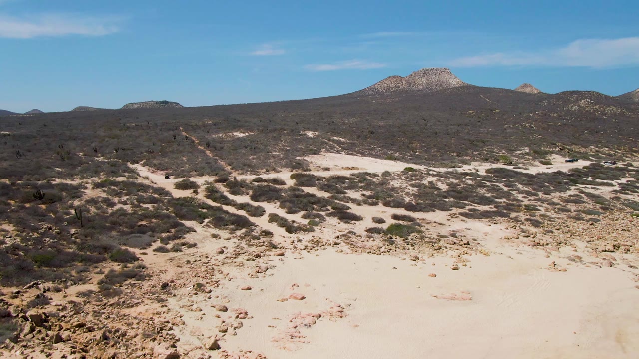 Aerial desert frame from Buzzards Beach with pale sand paths, dark low vegetation, rocky patches, a mountain ridge on the horizon, and no visible people.