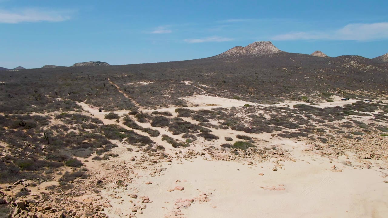 Wide aerial view over Buzzards Beach desert scrubland, with bright sand in the foreground, darker vegetation in the midground, distant rocky hills, and no visible people.
