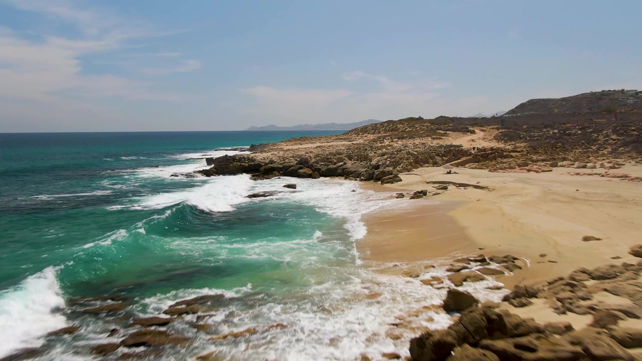 At Buzzards Beach in Baja California Sur, a foamy wave curls toward the sand while exposed rocks frame the lower edge and no people are visible.