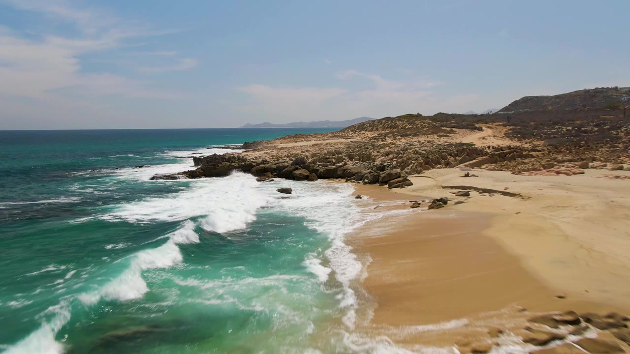 At Buzzards Beach in Baja California Sur, a broad sheet of white foam crosses the sandy cove with turquoise water offshore, rocky points, and no visible people.