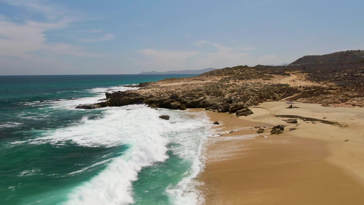 At Buzzards Beach in Baja California Sur, a larger surf line rolls along the rocky coast, with tan beach to the right and no visible people.
