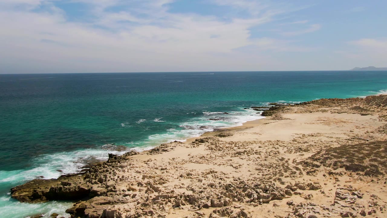 Aerial view of turquoise ocean breaking against a rocky sandy shoreline.