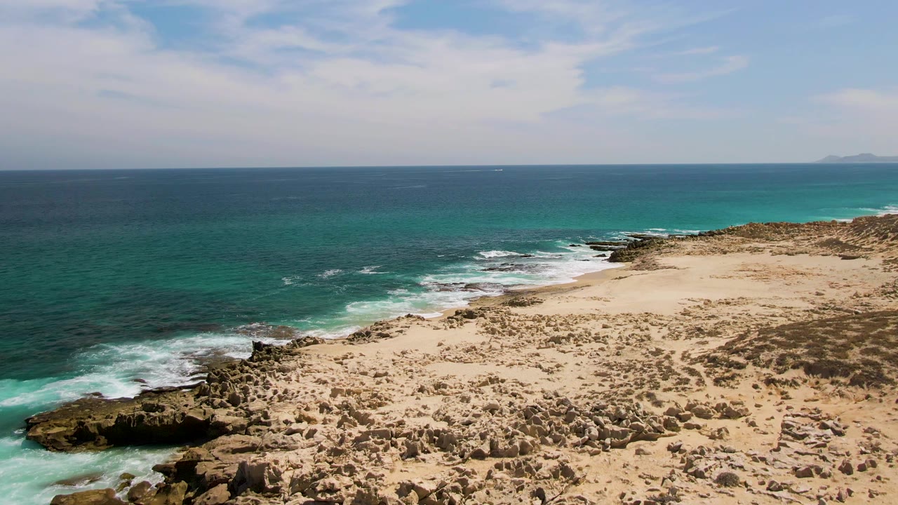 Aerial frame from Buzzards Beach with white waves wrapping around the left-side rocks, tan sandy ground across the foreground, blue ocean beyond, and no visible people.
