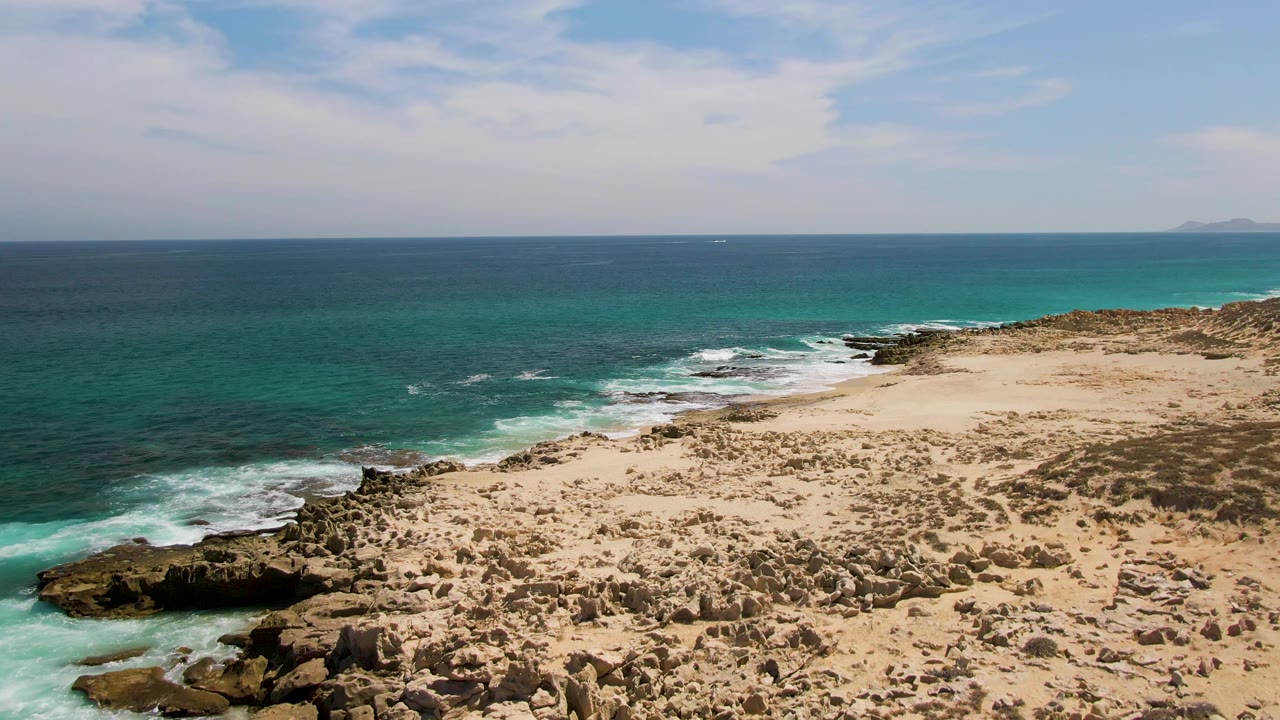 Aerial frame from Buzzards Beach showing a slightly farther view along the rocky sand bar, turquoise surf near shore, darker blue water offshore, faint distant land on the horizon, and no visible people.