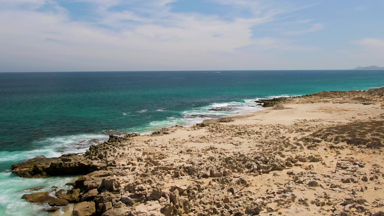 Aerial frame from Buzzards Beach with larger foreground rocks at the lower left, white water spreading into the sandy shoreline, clear turquoise shallows, open ocean, and no visible people.