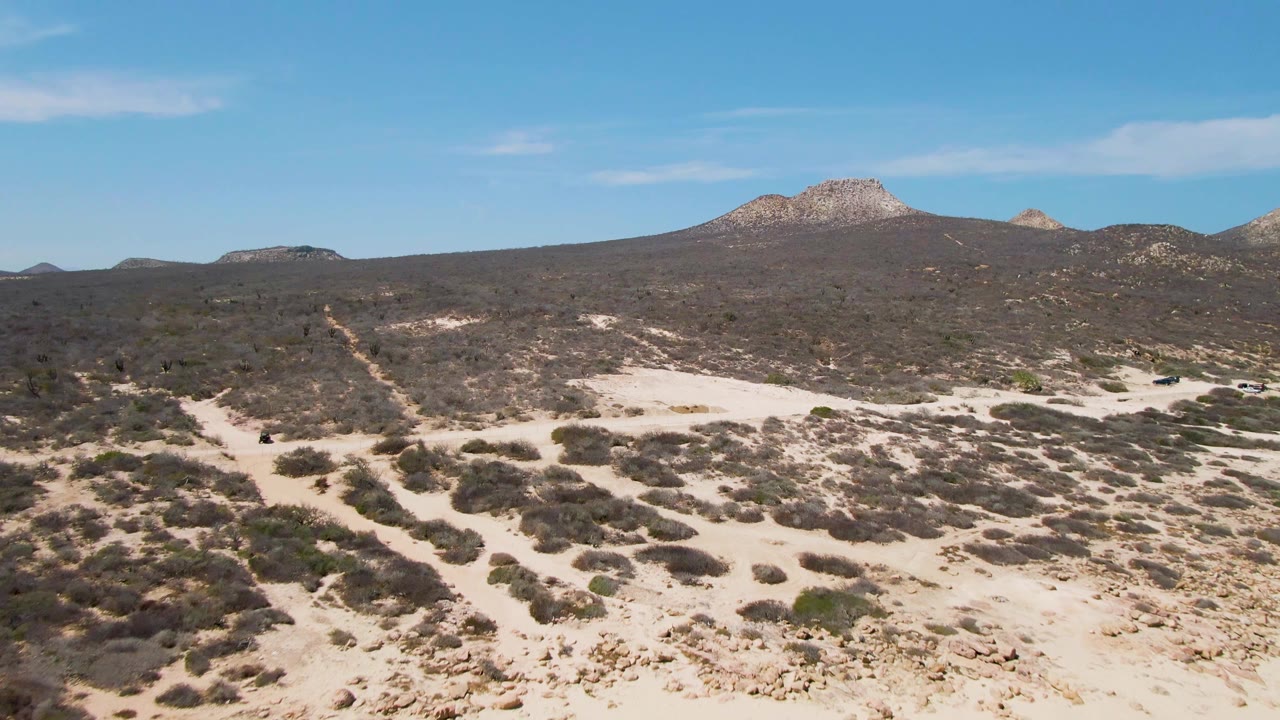 A small vehicle appears on the sandy track near Buzzards Beach, surrounded by low scrub, pale desert ground, rocky hills, and no visible people.