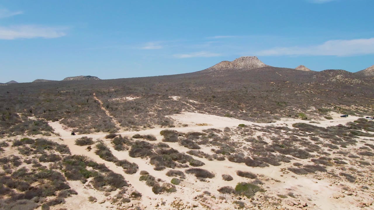 Mid-clip aerial view near Buzzards Beach showing the sandy road threaded through dark scrub patches toward rocky hills, with no visible people.