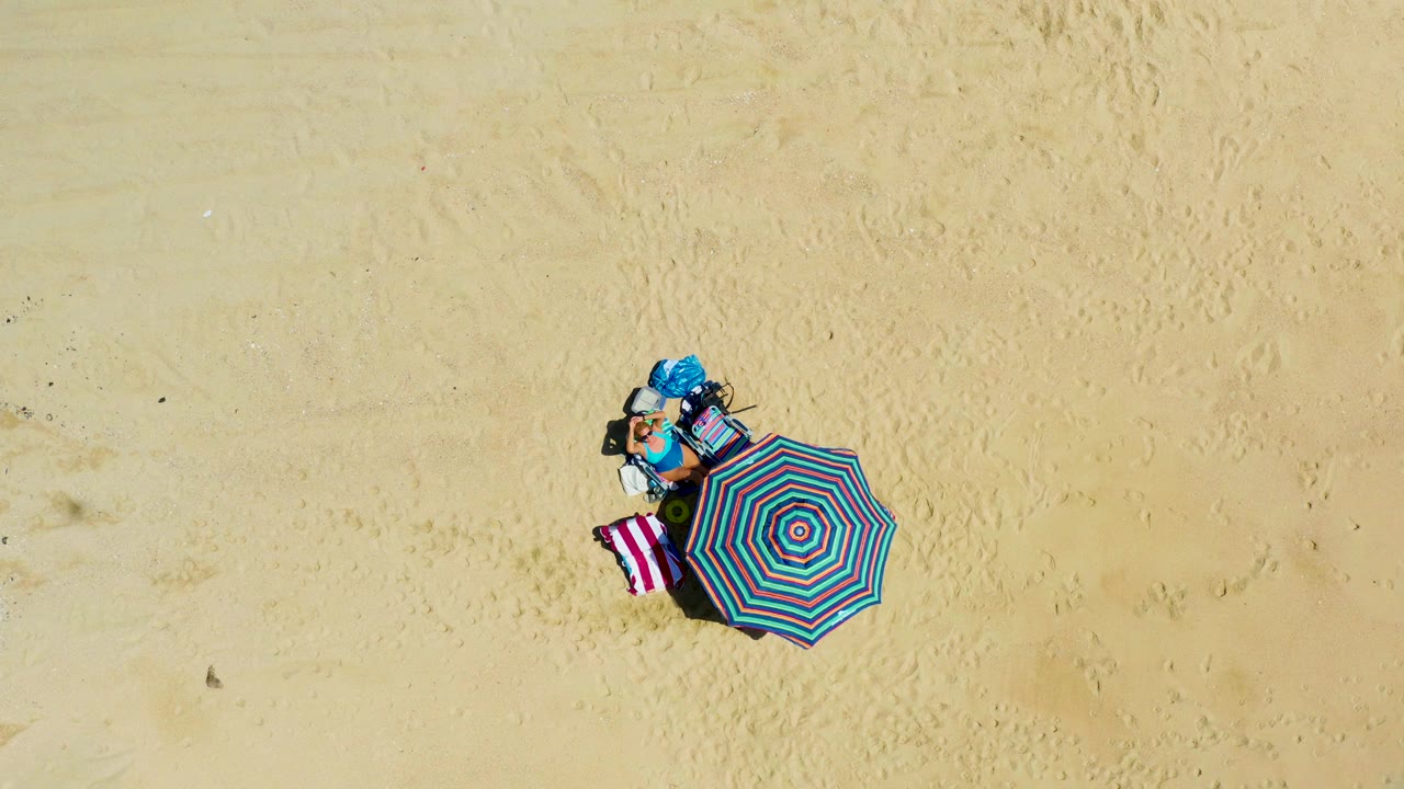 Overhead frame from La Laguna Beach Los Cabos, Mexico, showing a person beside a turquoise and pink striped umbrella with a pink and white towel on open sand.