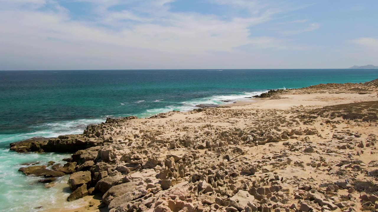 Rocky sandy coast beside turquoise ocean and pale sky.