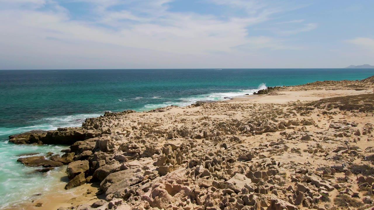 Early clip frame at Buzzards Beach showing a closer spread of sharp shoreline rocks, pale sand, white surf along the left cove, blue ocean, and no visible people.