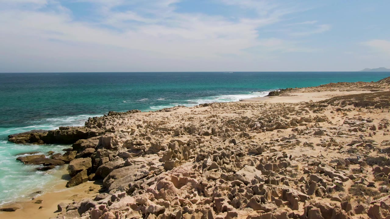 Buzzards Beach aerial frame with a wide field of eroded tan rock formations in the foreground, a narrow surf line at left, open turquoise sea, and no visible people.