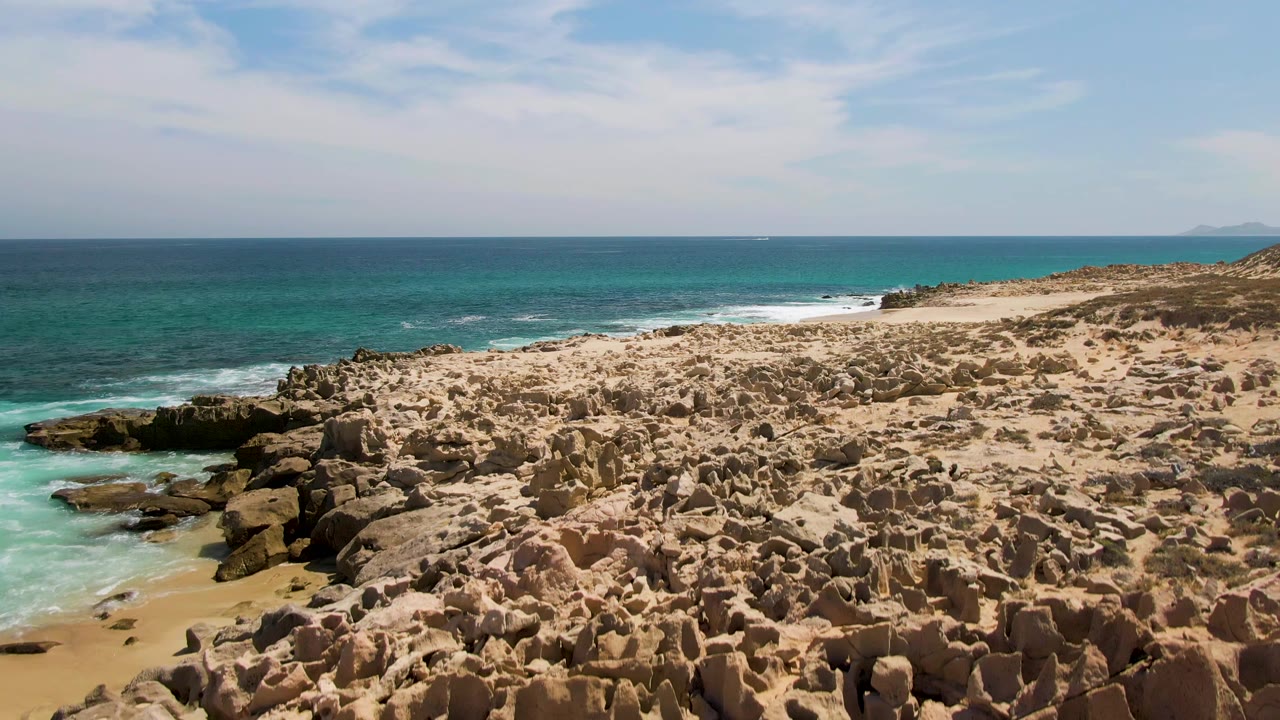 Mid-clip aerial view of Buzzards Beach, with foreground rock formations extending across the lower frame, turquoise water and foam to the left, sandy shore to the right, and no visible people.