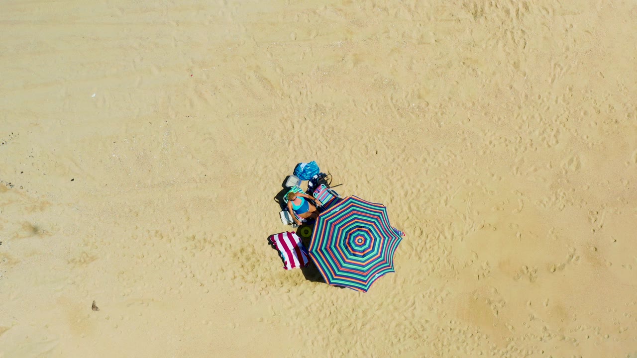 At 0:02, the overhead drone angle shows the striped umbrella beside the towel and beach gear on La Laguna Beach Los Cabos sand, with the person remaining near the setup.