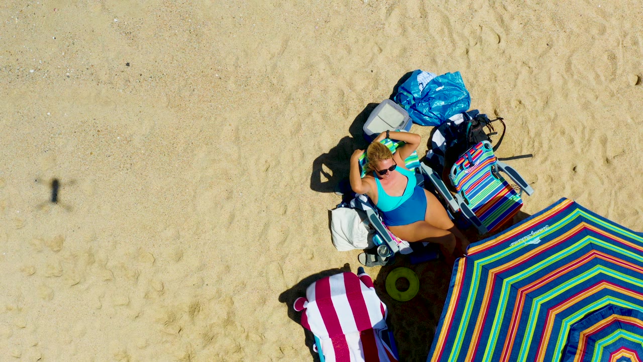 Overhead drone frame at La Laguna Beach Los Cabos showing a woman reclining on a beach chair beside a multicolored umbrella, beach bags, and a red-and-white towel on pale sand.