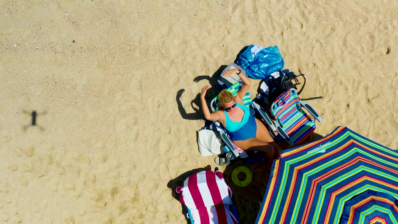Top-down view at La Laguna Beach Los Cabos with the woman raising a hand near her hair or sunglasses while lying beside the bright striped umbrella and packed beach items.