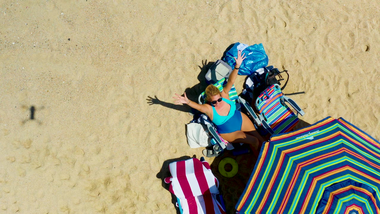Overhead frame at La Laguna Beach Los Cabos showing the woman with arms raised near her beach chair, with the striped umbrella, bags, towel, and long arm shadows visible on the sand.