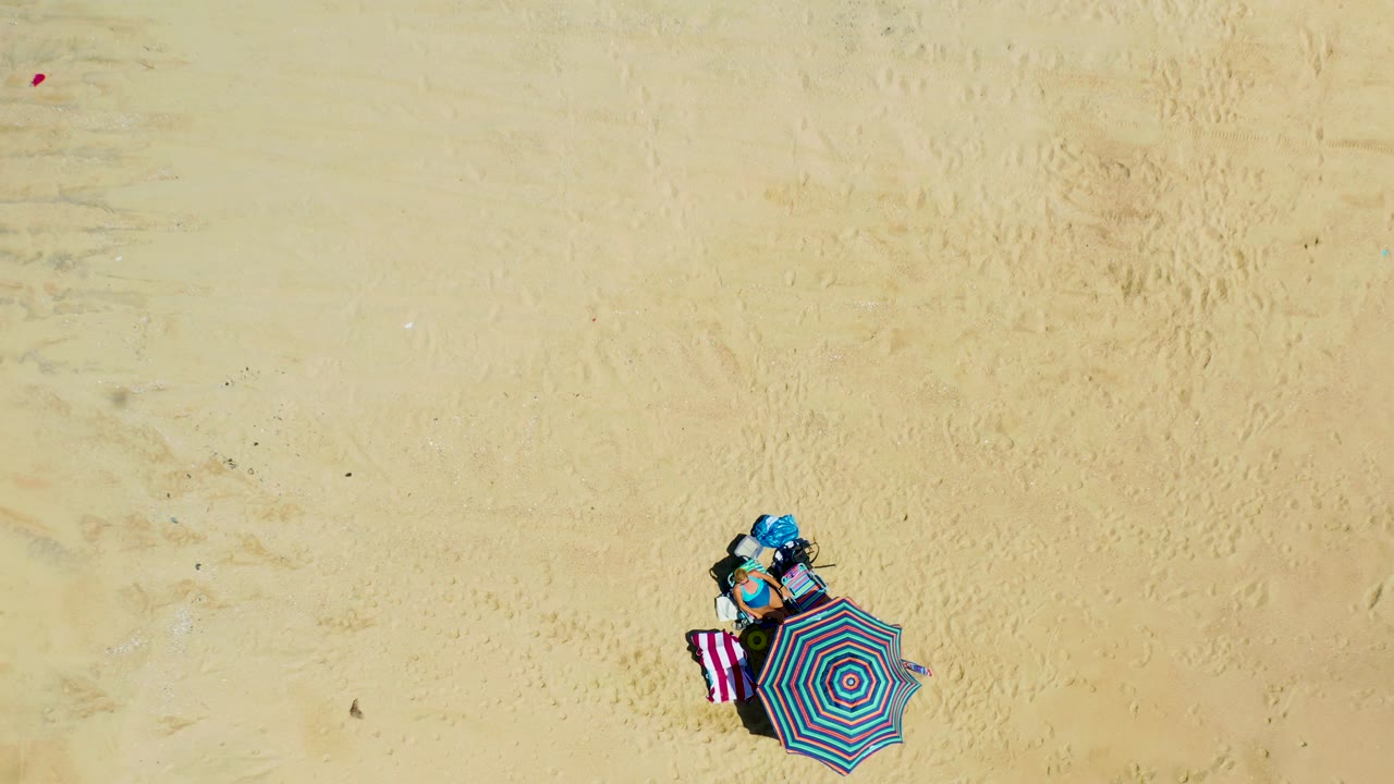 Direct overhead view at La Laguna Beach, Los Cabos, Mexico, with the striped umbrella and towel nearer the lower center, surrounded by pale sand tracks and no visible people.