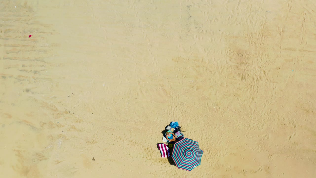 High overhead frame at La Laguna Beach, Los Cabos, Mexico, showing the compact umbrella and towel setup on the right side of a broad empty sand field, with no visible people.