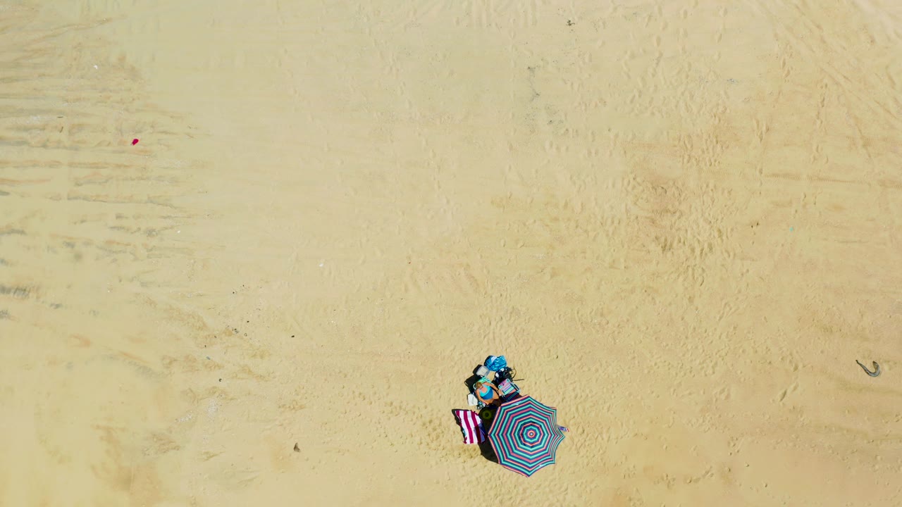 Overhead drone view at La Laguna Beach, Los Cabos, Mexico, with the umbrella and towel near the bottom edge, faint sand tracks across the frame, and no visible people.