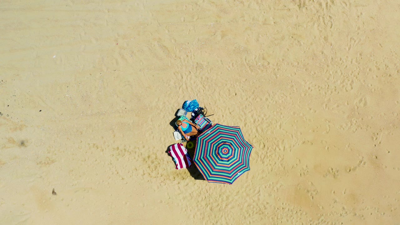 Overhead view of striped beach umbrella, towel, and blue bag on sand.