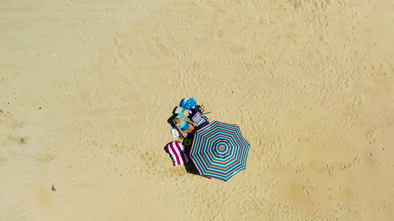 At 0:01, the top-down frame keeps the striped umbrella, towel, and blue item grouped on open sand at La Laguna Beach Los Cabos, Mexico, with no visible people.