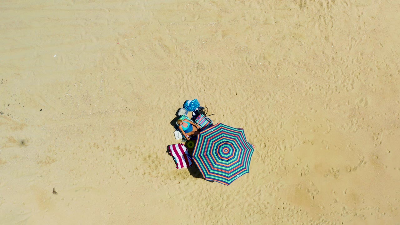At 0:02, the aerial view holds on the striped umbrella and towel while the blue bag remains near the upper side of the setup on the sandy beach in La Laguna Beach Los Cabos, Mexico, with no visible people.
