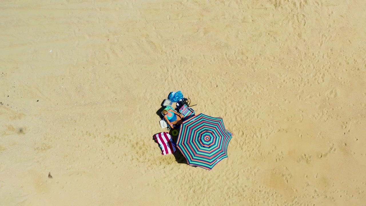 At 0:03, the top-down shot shows the striped umbrella and towel still fixed on pale sand at La Laguna Beach Los Cabos, Mexico, with the blue item less prominent and no visible people.