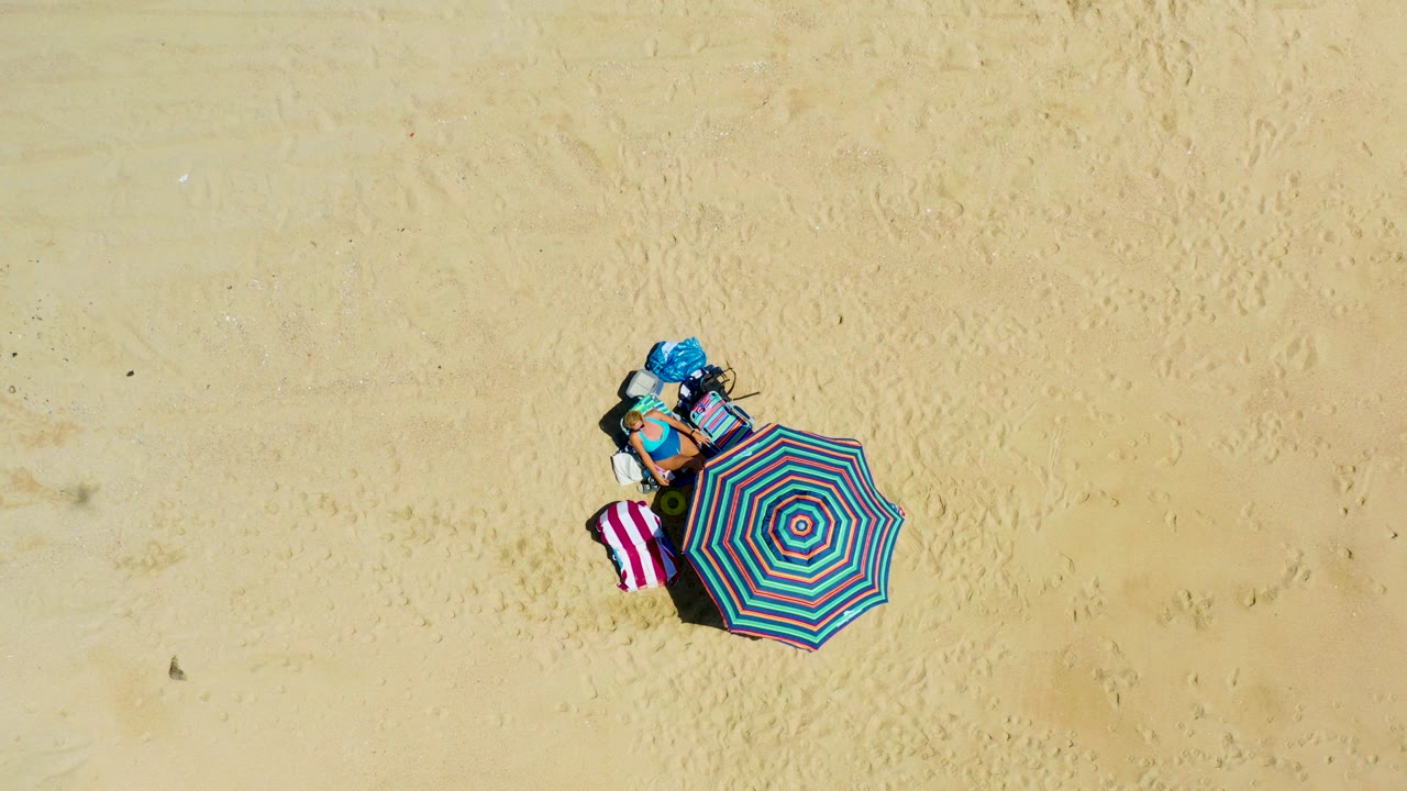 One second in, the La Laguna Beach overhead frame keeps the striped umbrella and towel centered low in the sandy composition, with no visible people.