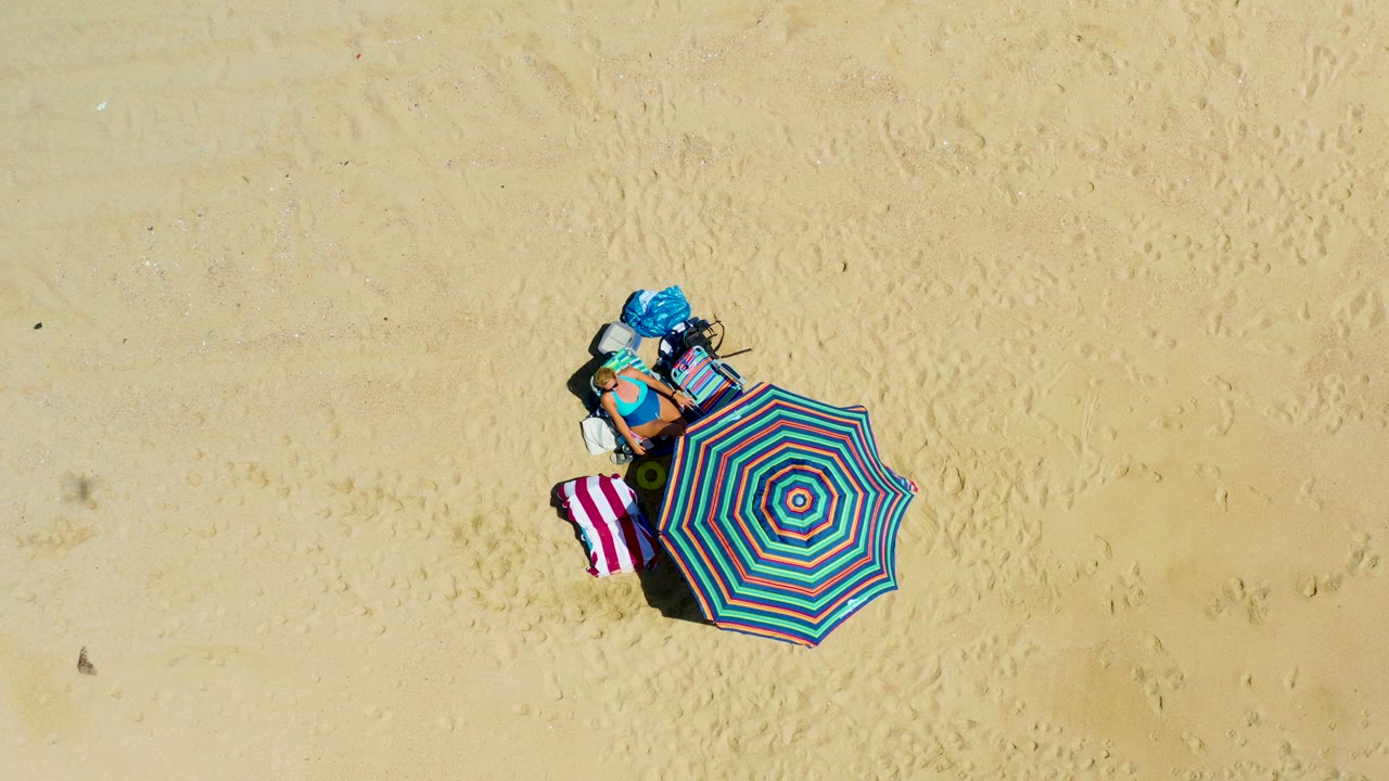 At 0:02, the La Laguna Beach drone view shows the umbrella, towel, and clustered belongings on pale sand as their shadows sit close beside them, with no visible people.