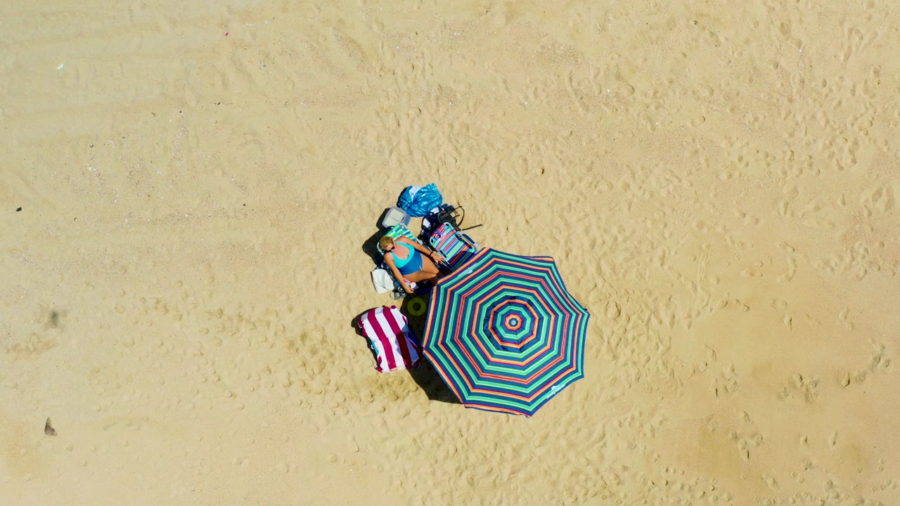 At 0:03, the same La Laguna Beach setup remains isolated in the lower-left area of the overhead frame, showing umbrella stripes, towel stripes, bags, and no visible people.