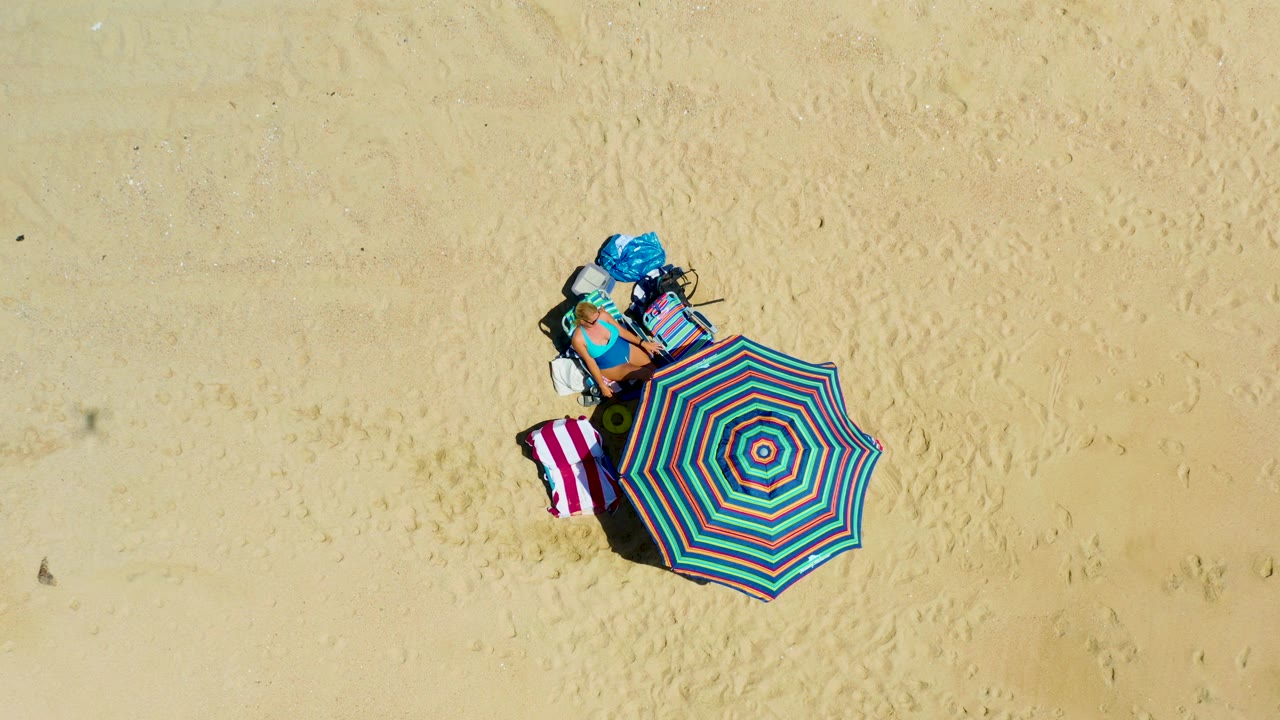 Overhead frame from La Laguna Beach Los Cabos, Mexico showing a colorful striped beach umbrella, towels, bags, and a seated person on pale sand.