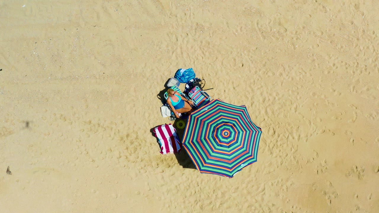 Striped beach umbrella, towels, bags, and person seen from above.
