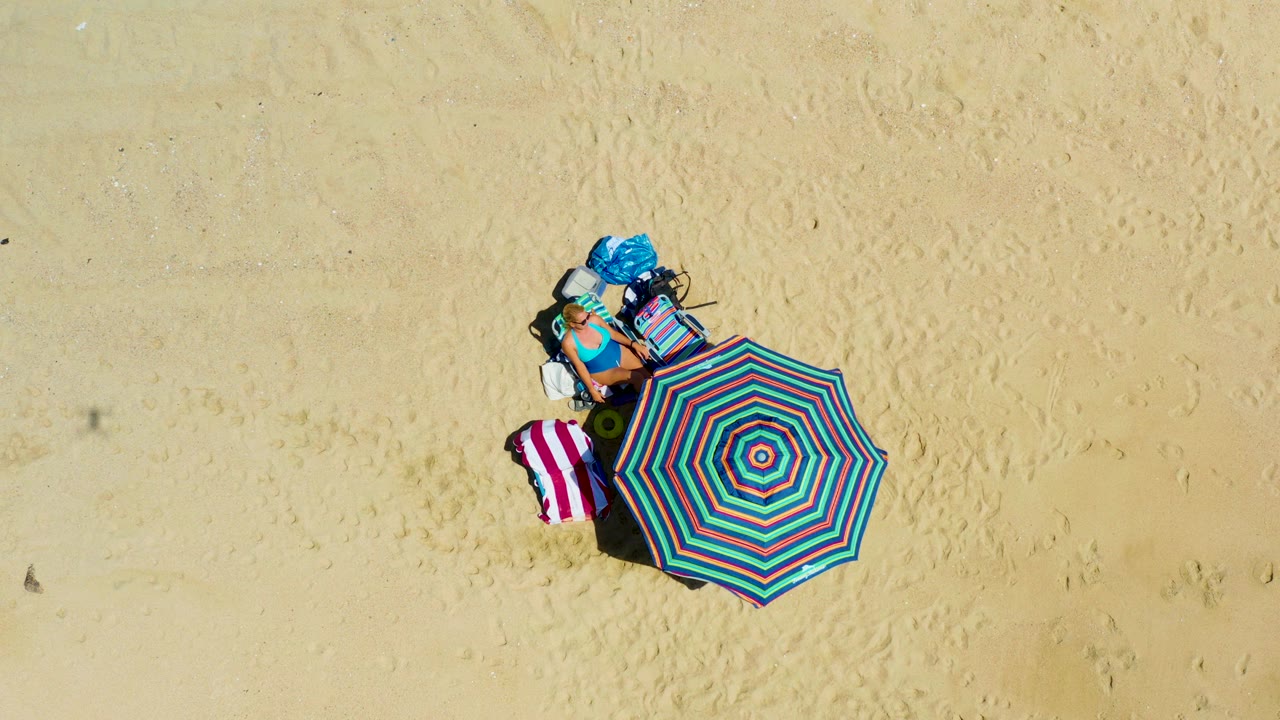 At 0:02, the aerial composition shows the turquoise and red striped umbrella with towels and bags arranged on pale sand at La Laguna Beach Los Cabos, Mexico.