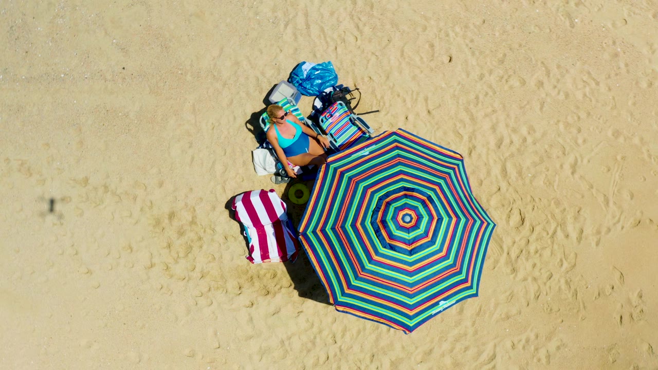 Overhead view at La Laguna Beach Los Cabos showing a woman in a blue swimsuit, a colorful striped umbrella, a towel, a chair, and bags on pale sand.