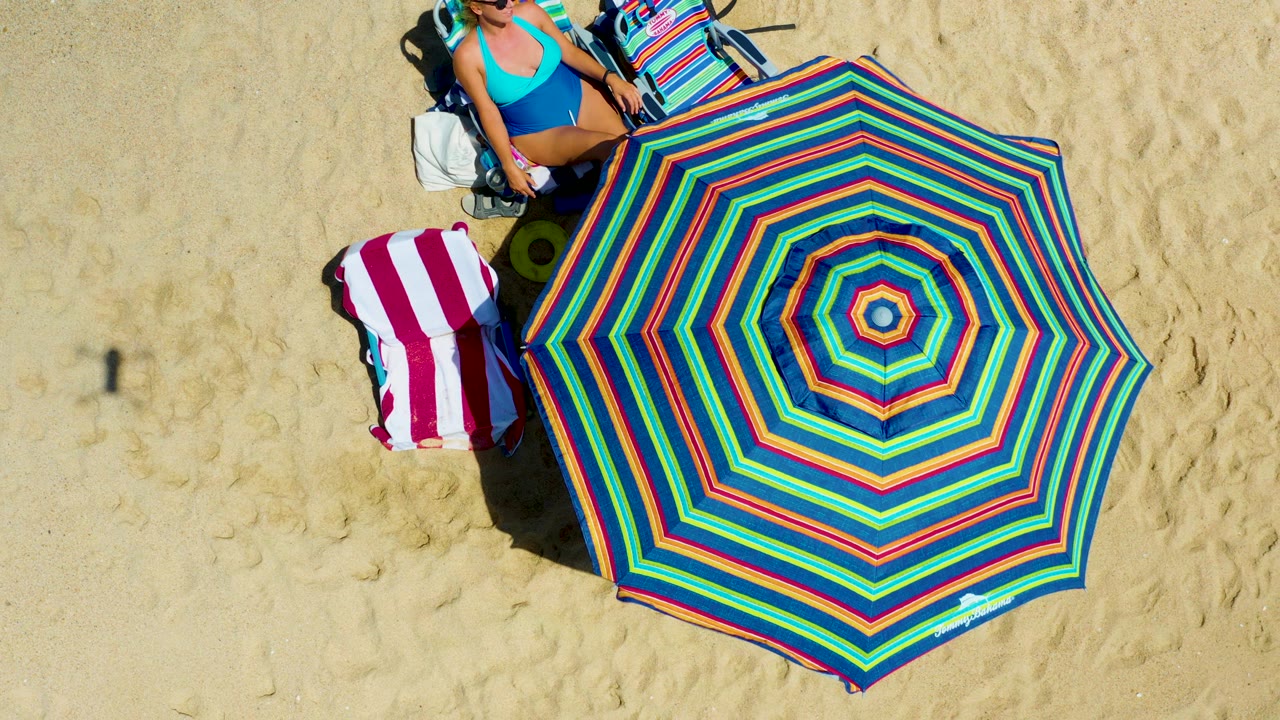 Overhead La Laguna Beach Los Cabos frame with the colorful umbrella dominating the lower right, the woman and chair at the top edge, and a striped towel on the sand.