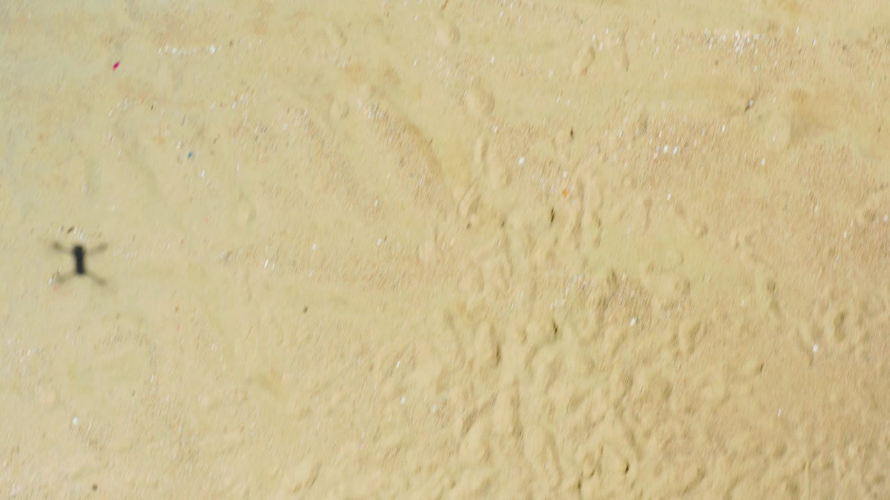At La Laguna Beach in Los Cabos, Mexico, an overhead frame shows open tan sand with a small drone-shaped shadow and the beach setup outside the visible area.