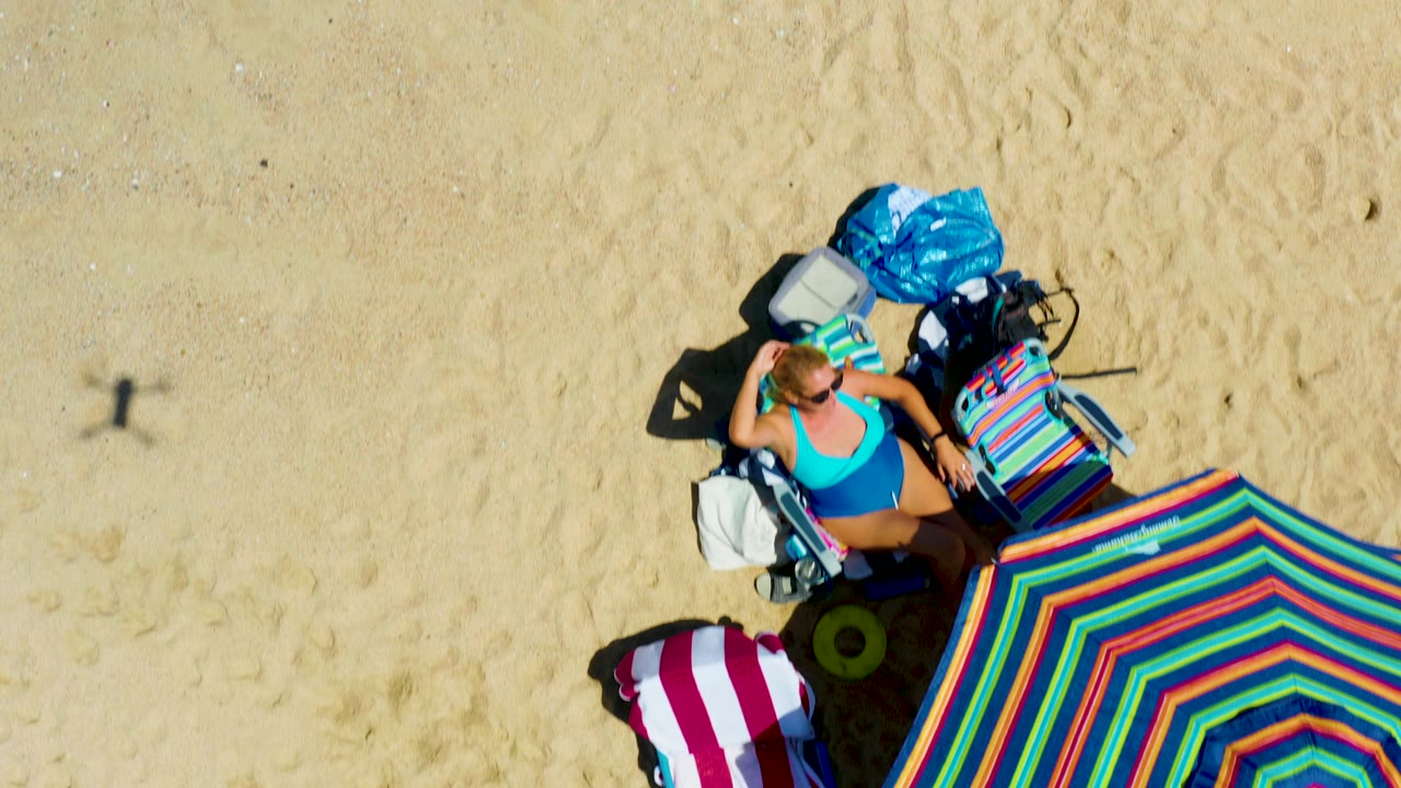 At La Laguna Beach in Los Cabos, Mexico, an overhead view shows a woman in a blue swimsuit on a lounge chair beside a colorful striped umbrella, towel, cooler, and bags.