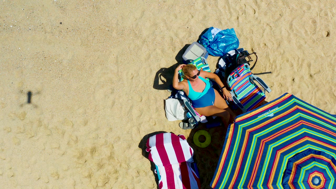 At La Laguna Beach in Los Cabos, Mexico, the overhead frame shows the woman near the top right of the umbrella, with the striped towel, cooler, bags, and hard shadows visible on the sand.