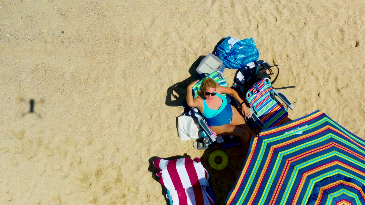 Overhead beach setup with woman, striped umbrella, towel, and gear.