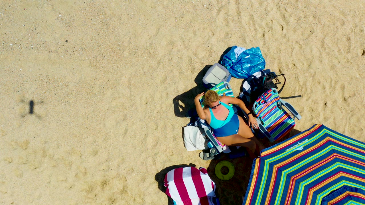 At La Laguna Beach in Los Cabos, a woman reclines on a beach lounge chair with one arm raised near her hair, next to a large striped umbrella and beach bags on pale sand.