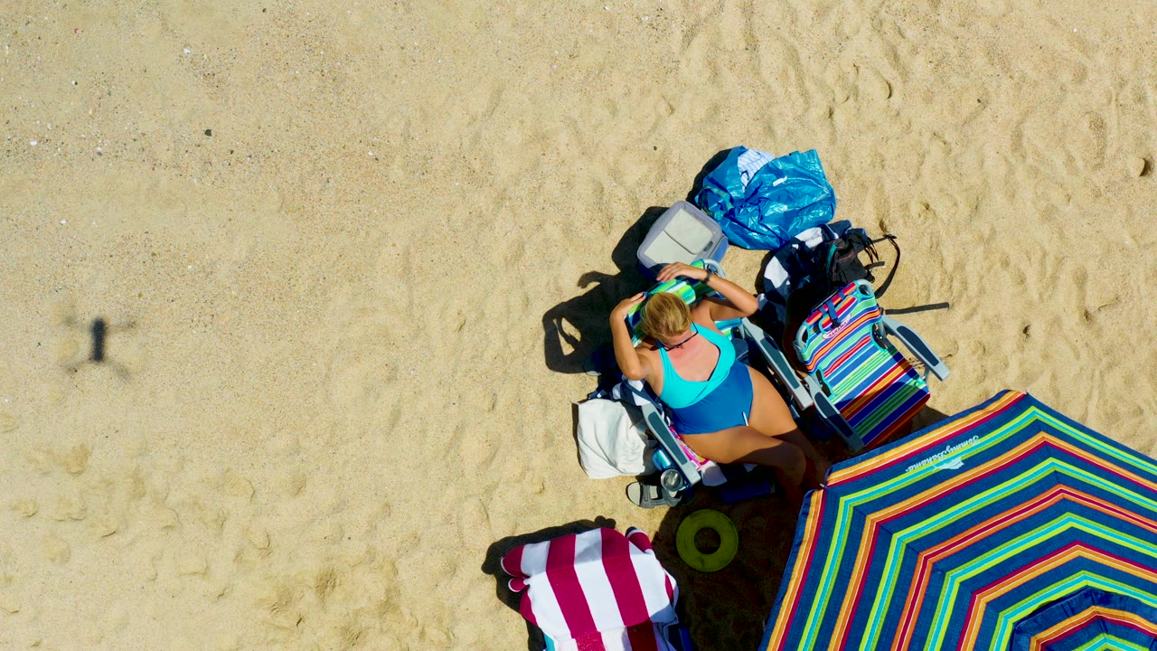 Woman on a lounge chair with beach bags, towels, and a large striped umbrella on sand.