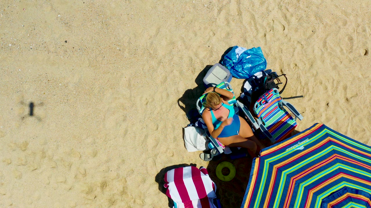 At 0:02, the woman remains on the lounge chair with her arm near her head, while the colorful striped umbrella fills the lower right of the La Laguna Beach frame.