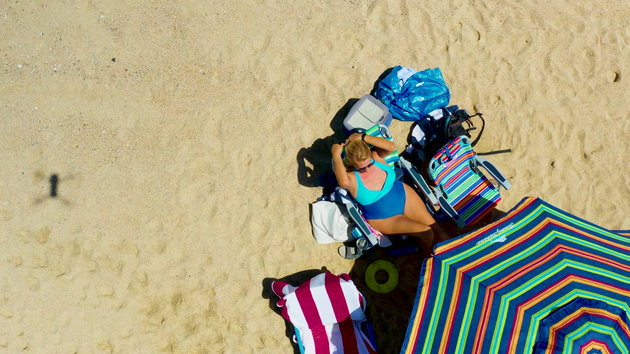 Top-down frame at La Laguna Beach Los Cabos, Mexico, showing a woman in a blue swimsuit on a beach chair beside a colorful striped umbrella, towel, bags, and open sand.